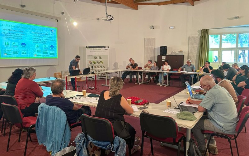 Des participants assis autour de tables en U &eacute;coutent un intervenant devant une projection sur les haies.