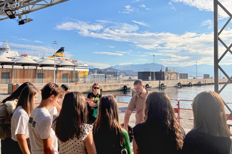 Un groupe de coll&eacute;giens &eacute;change avec un intervenant sur un stand en plein air, avec le port d'Ajaccio en arri&egrave;re-plan.